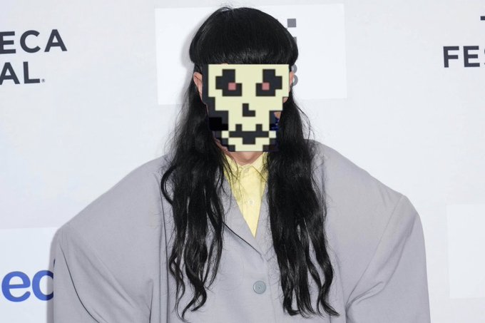 An edited photo of a person with long black hair wearing a grey coat, with their face obscured by a pixelated skull mask, standing in front of a Tribeca Festival backdrop.