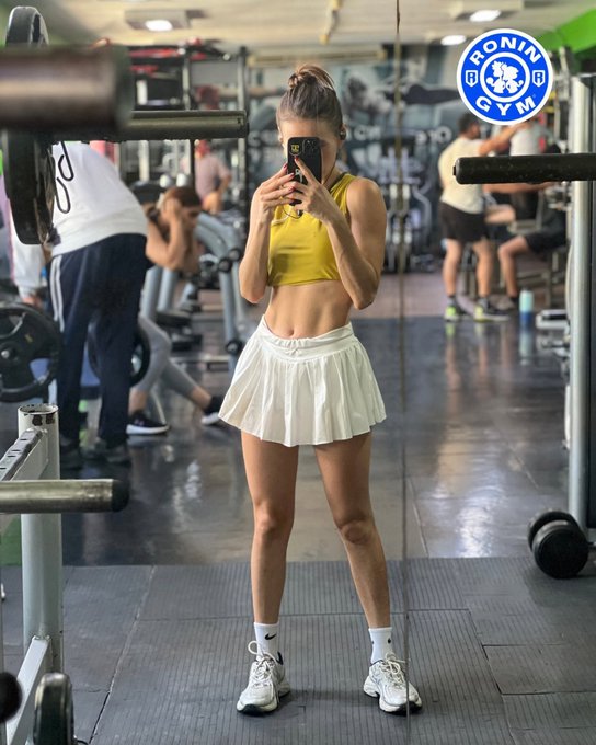 A woman in a yellow sports bra and white pleated skirt takes a mirror selfie with her phone at a gym, with a 'RONIN GYM' logo visible in the upper right.