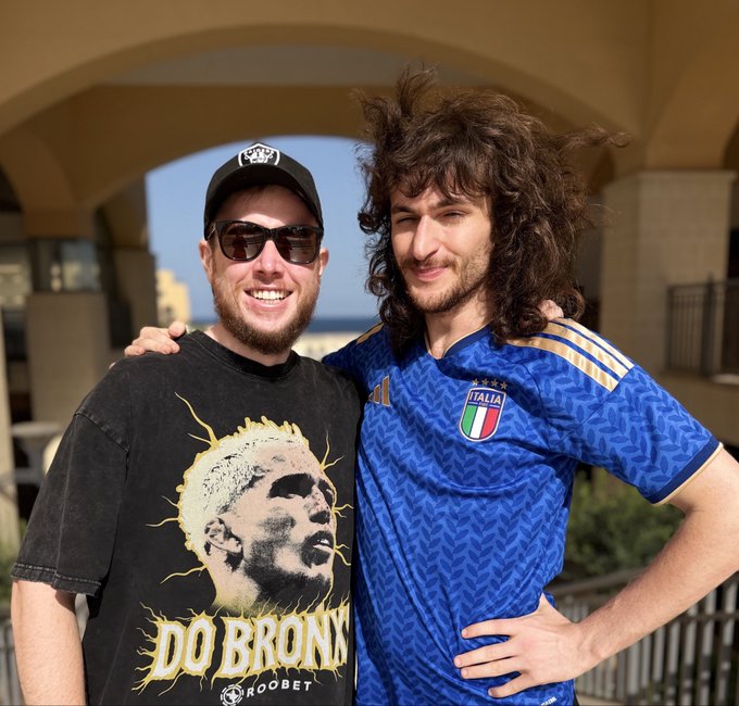 The caption could reference what the person is wearing or a feature of them, not focus on the event or the background. Here's a caption that meets the criteria:

A man with curly hair wearing a blue "ITALIA" jersey standing next to his friend in a black "DO BRONX" graphic t-shirt and cap.

The image shows two men posing together with arms around each other, with the visual elements (specifically the "DO BRONX" t-shirt) suggesting they may be fans or associates of MMA fighter Charles Oliveira, whose nickname is "Do Bronx." The "ITALIA" jersey worn by the curly-haired man adds a personal touch that contest participants might creatively incorporate into AI-generated versions of the photo.