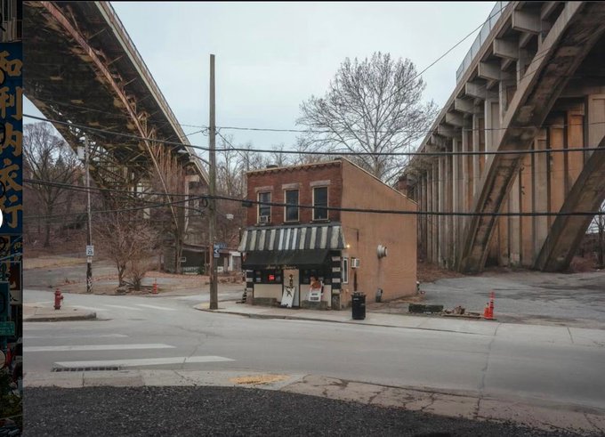 A photograph of a small brick building with a green awning situated underneath two large bridges or overpasses.
