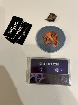 The image shows a collection of items, including two black and white cards with the words "ASYNC" and "SPIRITFLESH", a small circular item resembling a mask or pin, a mixtape, and some stickers. The objects are arranged on a table in front of a white wall.