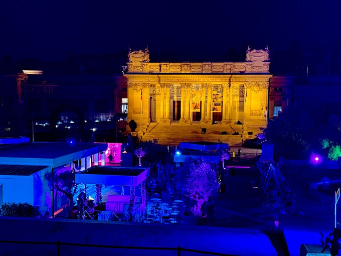 A grand historic building illuminated in golden light at night, with vibrant blue and purple lighting creating a colorful atmosphere in the foreground plaza and surrounding trees.