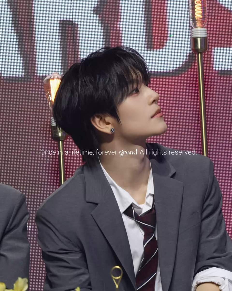 The image features a young man wearing a suit, tie, and earrings sitting at a table with his back turned towards the camera. The background of the photo includes a large screen displaying text that reads "260620 Eye-catching baby #XINLONG #鑫隆 #씬롱 파드라이브원 #ALPHADRIVEONE #ALD1". The man's attire and the presence of a large screen suggest that this image might be related to an event or gathering, possibly a fashion show or a promotional campaign for a brand.