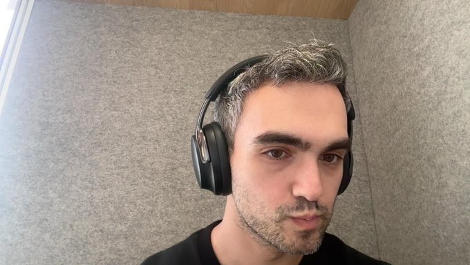 A close-up selfie of a man with short graying hair wearing black over-ear headphones in a room with gray sound-dampening walls.