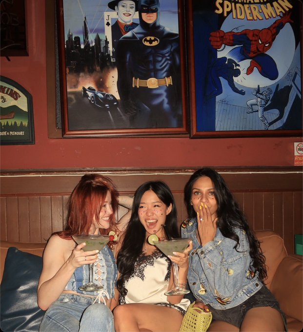 Three young women laugh and smile while holding margarita glasses on a leather couch. Behind them on a red wall hang framed posters featuring Batman (with a Joker playing card) and Spider-Man.