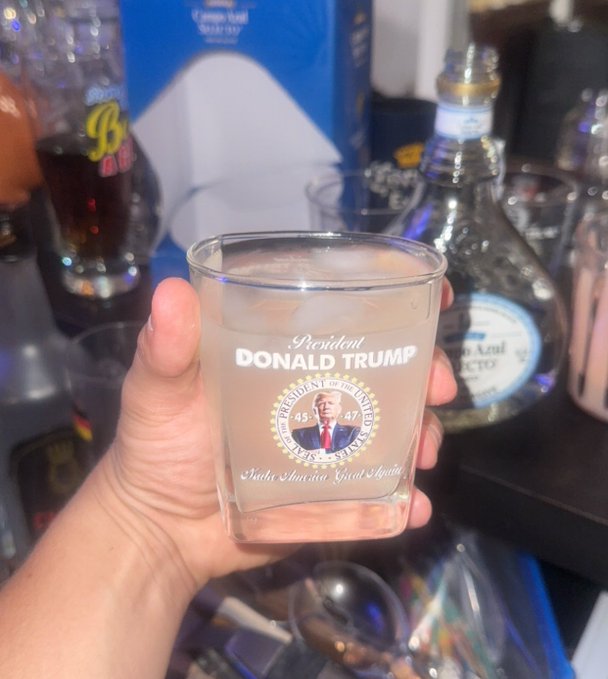 A hand holds a shot glass etched with 'Donald Trump, President of the United States, 45/47, Make America Great Again,' with liquor bottles visible in the background.
