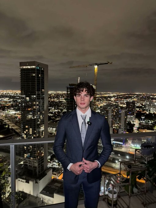 The image features a young man wearing a suit, standing on a balcony or deck at night with a city skyline visible behind him. He appears to be posing for a photo while looking directly into the camera. The caption "Jup mobile intern btw" is present in the image, which might be related to an event or occasion taking place in the city.