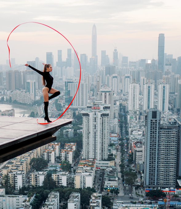 A woman in black stands on the edge of a high-rise building ledge swinging a red ribbon over a city skyline.
