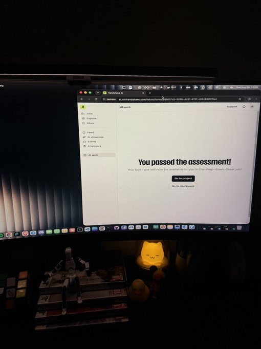 A computer monitor displays a screen with the bold text "You passed the assessment!" along with the message "This task type will now be available to you in the drop-down. Great job!" A small glowing yellow cat lamp and various figures sit on the desk below.