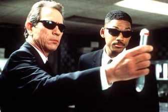 Two men in black suits and dark sunglasses — one older with gray hair, the other younger — stand in a corporate-style setting, both holding up small cylindrical objects while looking at them intently. The image evokes a "blind" or out-of-touch sensibility, fitting the post's commentary about traders whose bear-market tactics won't carry them through bullish conditions.