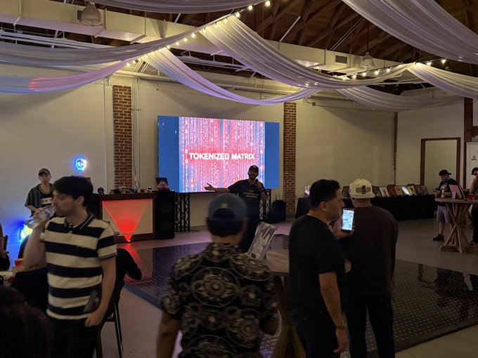 An indoor event venue with draped fabric ceilings and string lights, where attendees gather in front of a large screen displaying 'TOKENIZED MATRIX' in red text.