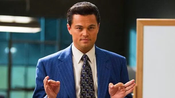 Leonardo DiCaprio, dressed in a blue pinstripe suit and patterned tie, stands in an office setting with both hands raised in a gesturing pose.