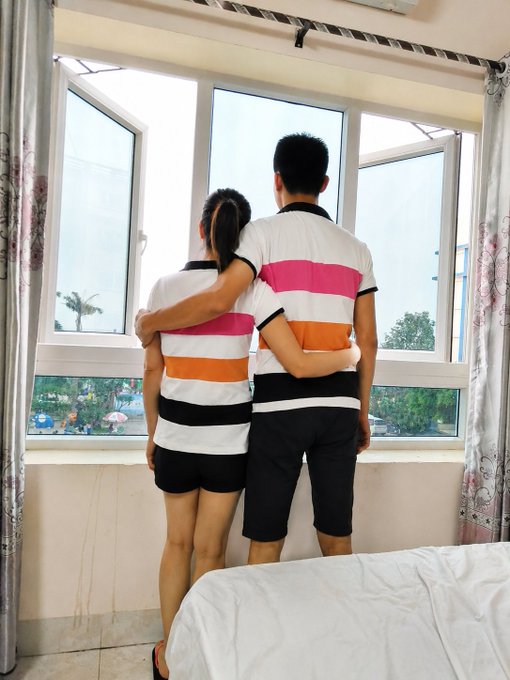 Two people wearing matching white, pink, orange, and black striped polo shirts embrace while looking out a window at a city view, suggesting a family photo or couple's moment.