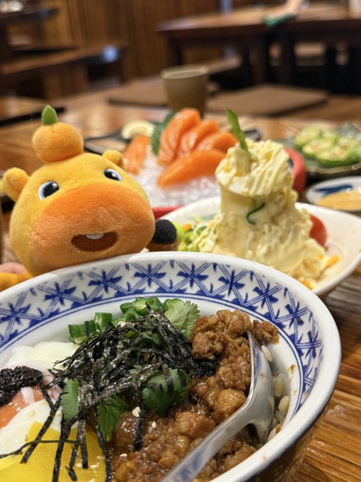 A close-up photo of a Japanese meal featuring a rice bowl topped with seaweed, meat, and egg, alongside a yellow plush toy, salmon sashimi, and potato salad in a restaurant setting.