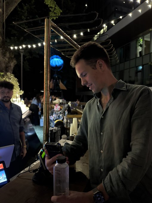 A man wearing a green shirt and cap stands at a bar at night, holding a clear bottle while looking down at his phone, with string lights overhead.