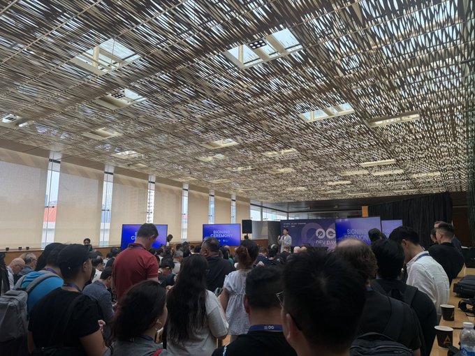 A crowded conference room with an intricate wooden slat ceiling where attendees gather for what seems to be the Singapore FinTech Festival, with screens showing 'OCBC' and 'SONIC BLOCKCHAIN'.