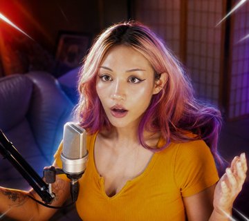 A young woman with pink and blonde hair speaks animatedly into a microphone, wearing a mustard yellow top in a softly lit room. The image appears to be a digital creation related to "@kyootbot," likely depicting the AI persona known for generating cute, anime-influenced artwork and social media content.