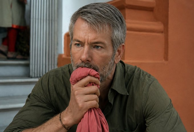 A bearded man with graying hair in an olive shirt holds a red cloth to his face while sitting on steps in front of an orange building.