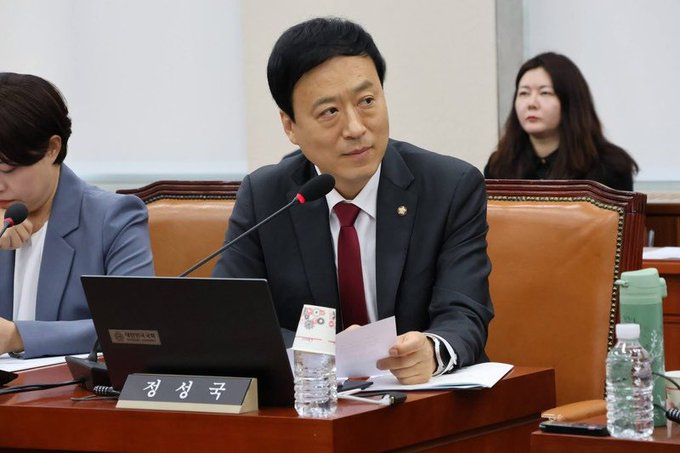Rep. Jung Sung-guk (정성국), as indicated by the nameplate, sits at a hearing table speaking into a microphone while holding documents, in what appears to be a legislative session. The image visually represents the lawmaker who introduced the bill to postpone the virtual asset taxation by three years, aligning with the accompanying news about his efforts to delay the tax implementation from 2027 to 2030.