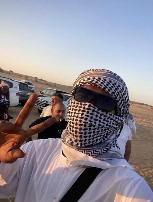 A person wearing a black and white checkered keffiyeh and sunglasses takes a selfie in a desert setting, making a peace sign while other men and parked vehicles are visible in the background.