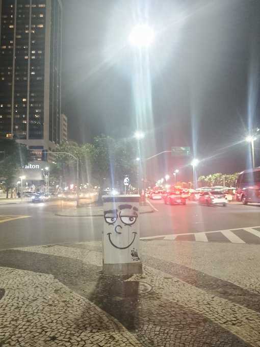 A nighttime street scene featuring a small utility box with a cartoon face drawn on it, standing on a patterned sidewalk at a crosswalk with cars passing by in the background.