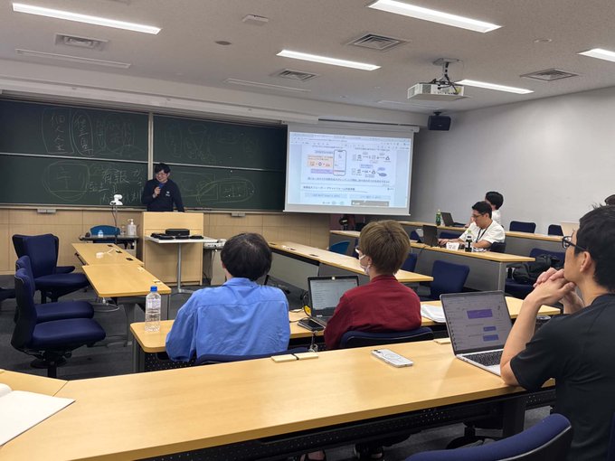 The image shows a classroom setting with two people sitting at desks, one of whom appears to be giving a presentation or lecture. The other person has their laptop open in front of them, possibly taking notes or following along with the content being presented. There are several chairs and desks arranged around the room, indicating that it is a typical classroom environment for educational purposes. The post about the lecture at Keio Business School suggests that this scene takes place within an academic setting where students can learn from experts in their respective fields.