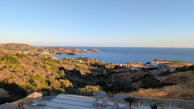 A scenic coastal landscape at sunset showing a bay with hills, a resort area with lounge chairs and umbrellas, and a few buildings overlooking the water.