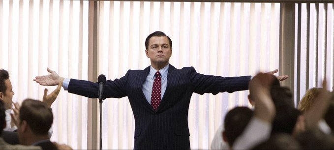 A man in a dark pinstripe suit and red tie stands confidently with arms outstretched before an applauding crowd, with vertical blinds visible behind him. The image complements the post by visually conveying the discipline, confidence, and patient expertise the author claims are essential to consistent trading profitability.