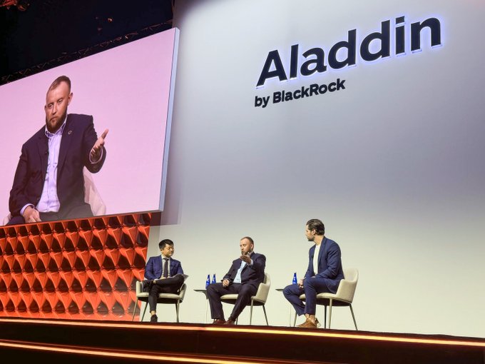 Panel discussion on stage at an 'Aladdin by BlackRock' event, featuring three men seated in white chairs with a large screen displaying one of the speakers.