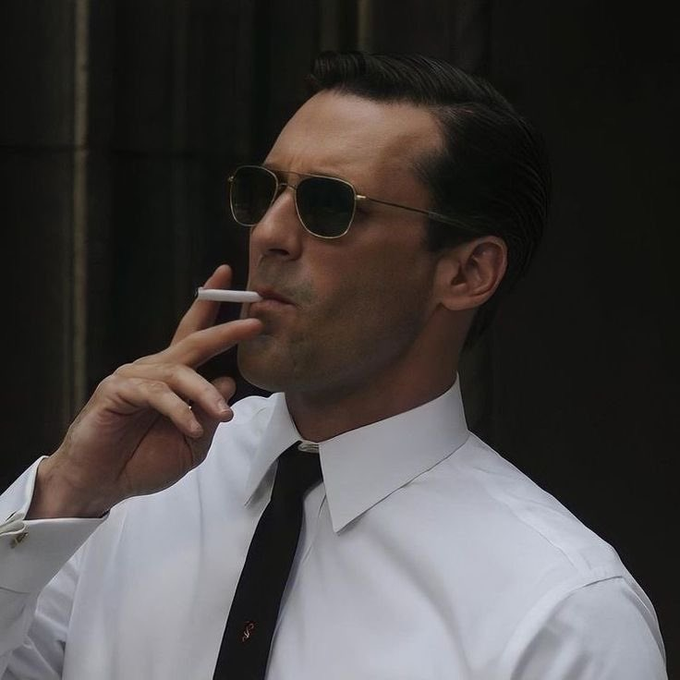 A man in a white shirt, black tie, and gold-rimmed sunglasses smoking a cigarette, styled to resemble a Don Draper aesthetic.