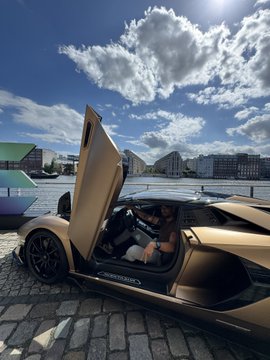 A bronze Lamborghini Aventador with its scissor doors open is parked on a cobblestone waterfront, with a person seated in the driver's seat.