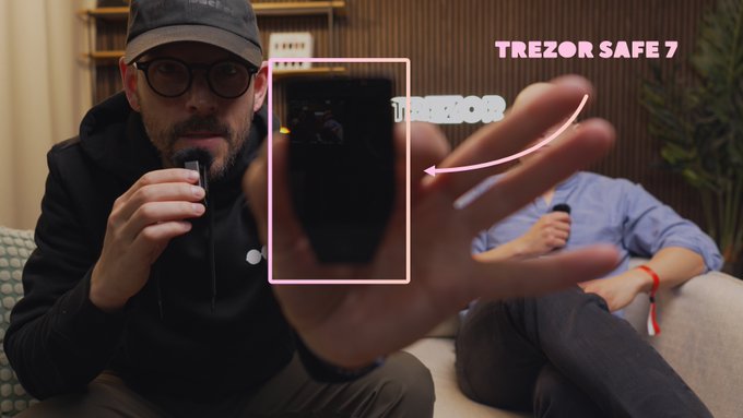 A man wearing a cap and glasses holding up a Trezor Safe 7 hardware wallet device, with the text 'TREZOR SAFE 7' displayed in pink on the right side.