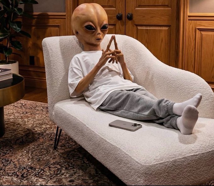 An alien figure wearing a white t-shirt and gray sweatpants lounges on a white chaise lounge, examining its fingers with a smartphone resting nearby. The image humorously suggests that with the right idea (prompt), anyone—even an extraterrestrial—could be on the verge of building a million-dollar app.