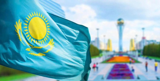 The flag of Kazakhstan waves in the foreground with the Baiterek monument and a city park scene.