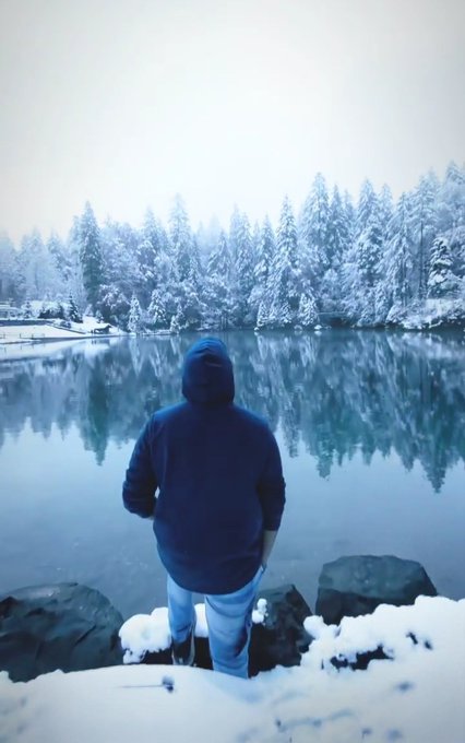 A person in a dark blue hoodie and light pants standing with their back to the camera, looking out over a calm lake surrounded by snow-covered pine trees in a wintery landscape.