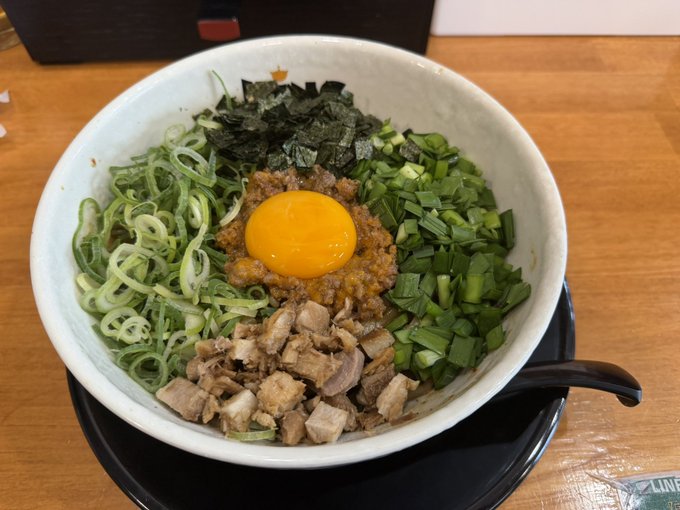 A bowl of Japanese-style noodle dish (possibly mazemen or abura soba) topped with a raw egg yolk, minced meat, sliced chashu pork, chopped green onions, nori seaweed, and leeks, served on a wooden table.
