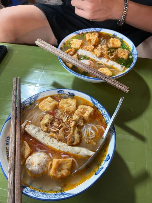 Two bowls of Vietnamese noodle soup (bún) with fried tofu, chả (fish cake), and quẩy served on a green table with chopsticks, one bowl with a spoon.