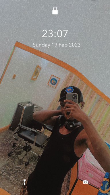A man wearing a black tank top, cap, and headphones takes a mirror selfie, with the phone screen showing the time 23:07 and date Sunday 19 Feb 2023.