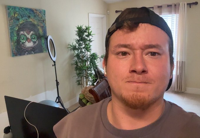 The image features a man wearing a baseball cap, sitting in front of a laptop computer with his face partially visible. There are two potted plants in the room - one near the left side and another towards the right side. The background includes a painting on the wall above the man's head and a window next to it. A caption below the image reads "No Haircut until Bitcoin goes over $100k again".