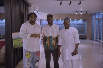 Three men pose together at an indoor event space in Abuja, Nigeria, with the center figure holding a colorful gift bag, capturing a moment from the recent Arc community meetup organized by local lead Jerry Ojumah.