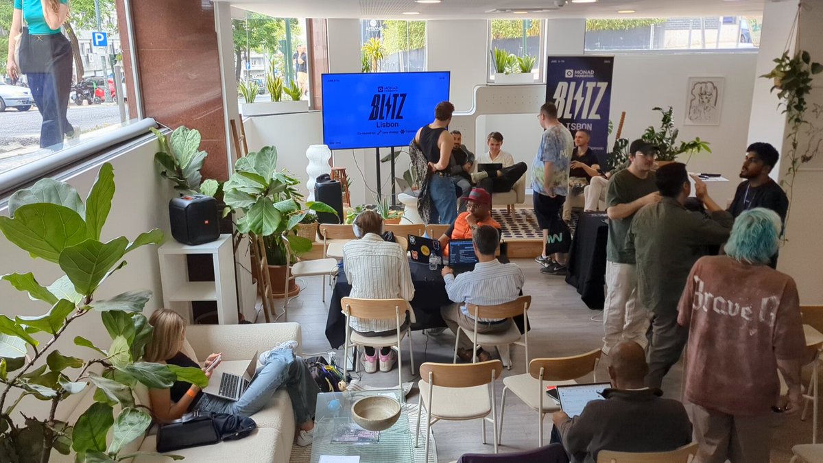 The image shows a group of people gathered together in an open room filled with furniture such as couches and chairs. There are potted plants placed throughout the space, adding greenery to the environment. A large TV screen can be seen on one side of the room, likely used for presentations or discussions. The attendees appear engaged in conversation, possibly discussing a project related to Monad Hacking at the @monad Blitz event happening today in Lisbon.