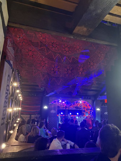 A crowded, dimly lit venue features a stage in the background illuminated with blue and red lights, with a decorative red crocheted canopy hanging from the wooden ceiling above the audience.