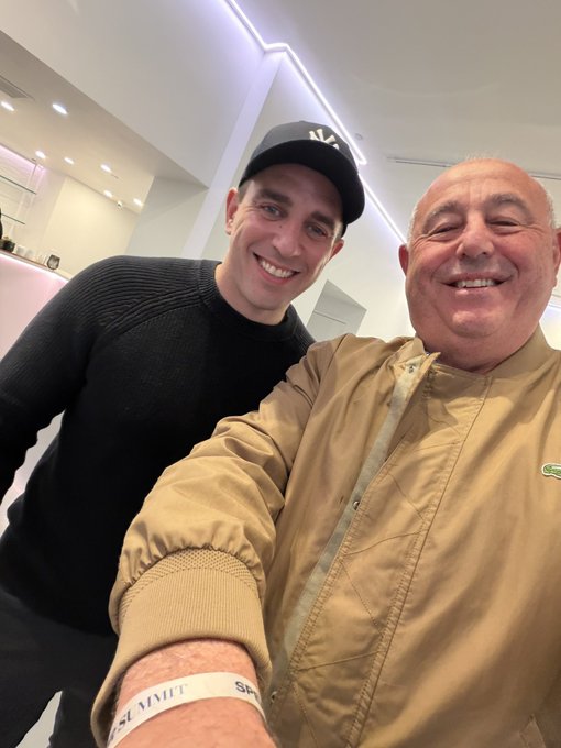 A selfie of two smiling men; one is younger, wearing a black cap and black sweater, while the older man on the right wears a tan Lacoste jacket.