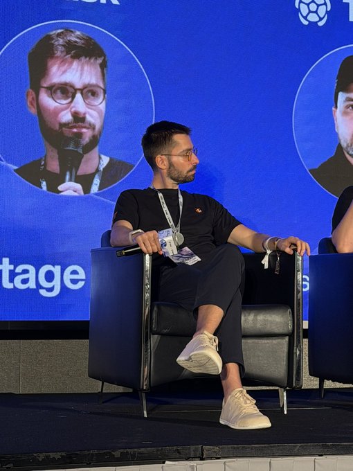 A man with glasses and a beard seated in a black armchair on stage at a conference, holding a microphone, with screens displaying his image behind him.