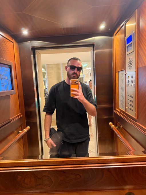 A bearded man in sunglasses takes a mirror selfie in an elegant wood-paneled elevator, capturing a stylish "Gm" greeting to his followers as he starts his day in style.