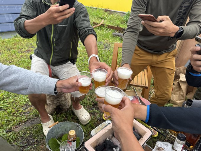 A group of friends clink plastic cups of beer in a toast outdoors, celebrating a gathering at a countryside home surrounded by mountains and sea, with a cooler of drinks and bottles nearby. One person wears a Mont-Bell jacket and Crocs, capturing the casual, joyful vibe that matches the post's invitation to revisit the scenic spot with @fukushunosuke and "shu."