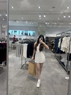 A mirror selfie taken inside a clothing store shows a woman wearing a short white dress and white sneakers while carrying a tan handbag.