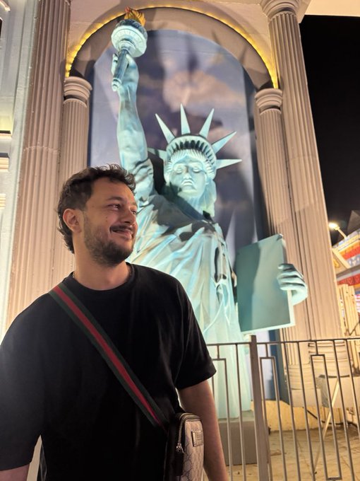 A man wearing a black t-shirt with a Gucci crossbody strap standing in front of a large replica Statue of Liberty at a Las Vegas casino at night.