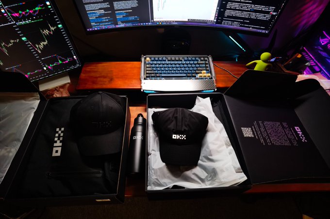 An OKX-branded merchandise setup on a wooden desk including a black cap, water bottle, beanie, and t-shirt in packaging, displayed in front of a multi-monitor trading workstation showing financial charts.