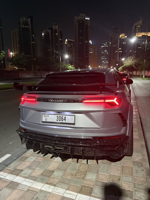 A silver Mansory-tuned Lamborghini Urus, displaying a Dubai license plate "U 3064," is parked on a city street at night with an illuminated skyline in the background. The "GN" caption likely refers to a username or tag associated with the owner showcasing their luxury modified vehicle.